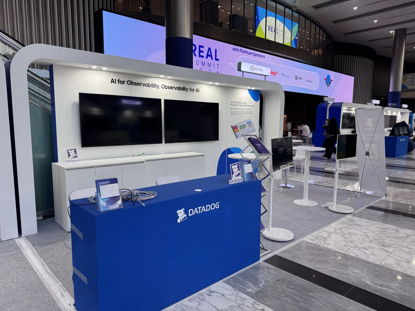 REAL Summit 2026 Datadog 부스 정면 — 파란 카운터, 벽면 TV 두 대와 슬로건, 오른쪽 스탠드형 디스플레이와 X배너